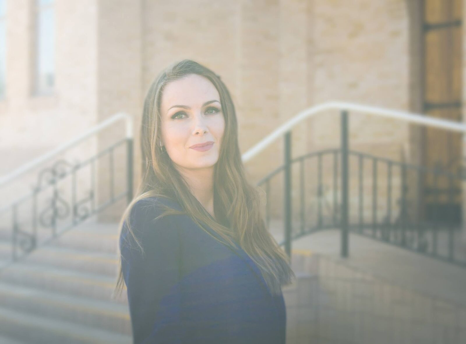 Portrait of a woman with long hair in a navy top, outdoors in front of stone steps with iron railings
